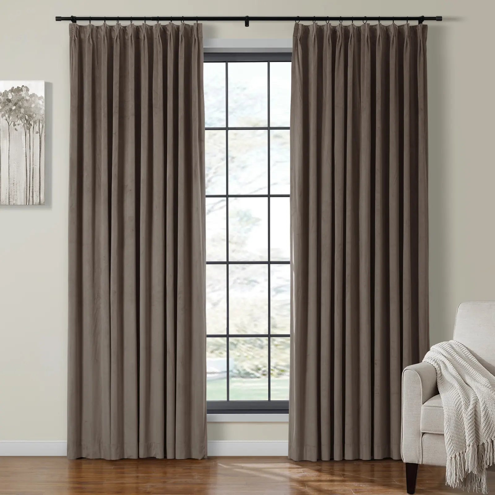 Plush taupe brown velvet pleated curtains hanging from a black rod frame a multi-pane window, beside an abstract gray tree artwork and a cream armchair with a fringed throw blanket
