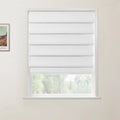 Framed print by Vincent van Gogh depicting a vibrant rural landscape with a red-roofed building and colorful fields hangs on a light beige wall beside a window fitted with a white layered fabric Roman blind that partially reveals a blurry green outdoor landscape