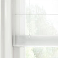 Photograph of a sheer white layered window blind featuring a subtle horizontal weave and thin control cords allowing diffuse light from the blurred green foliage outside