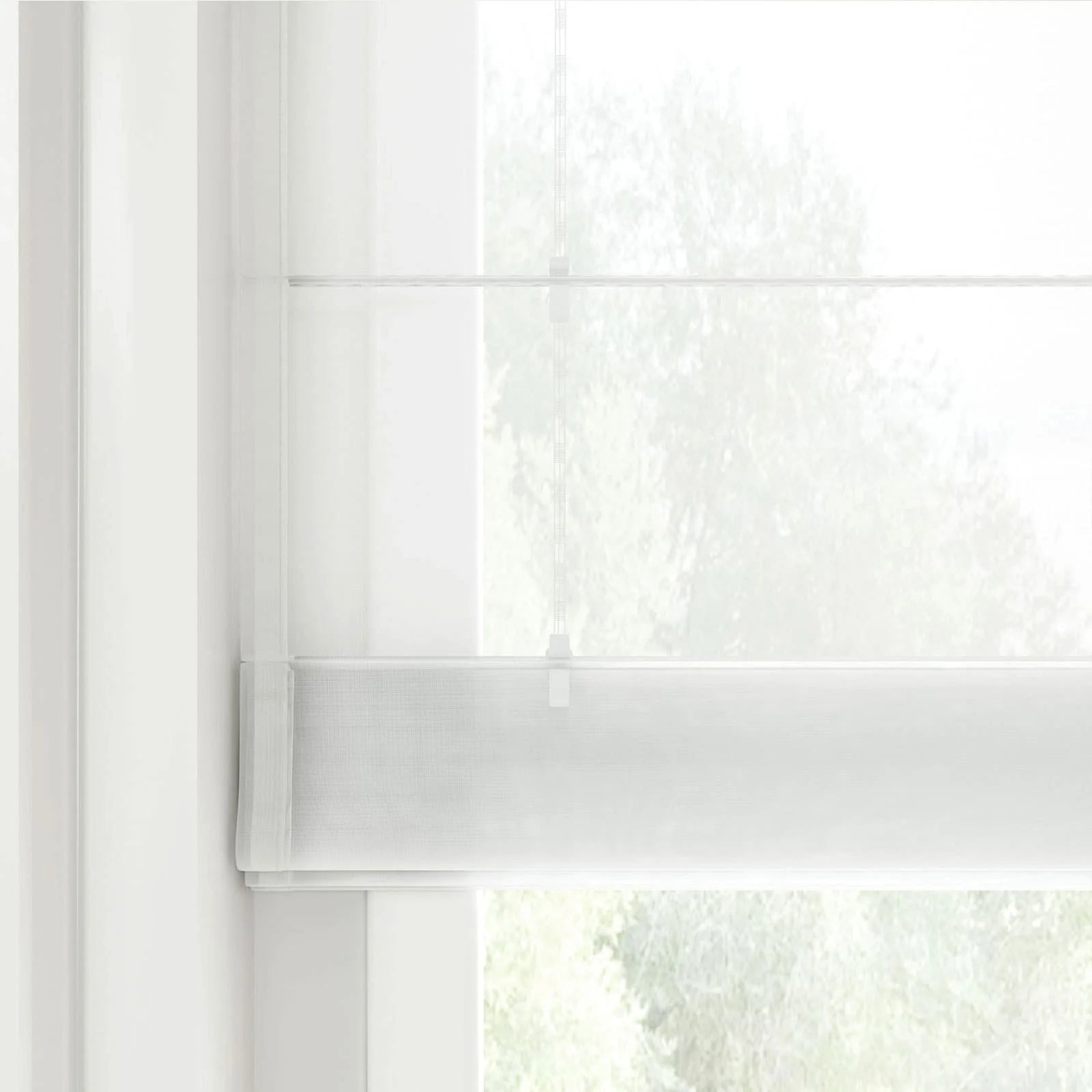 Photograph of a sheer white layered window blind featuring a subtle horizontal weave and thin control cords allowing diffuse light from the blurred green foliage outside