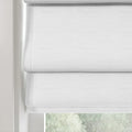 A white linen textured Roman blind with soft stacked folds partially revealing blurred green foliage outside a window