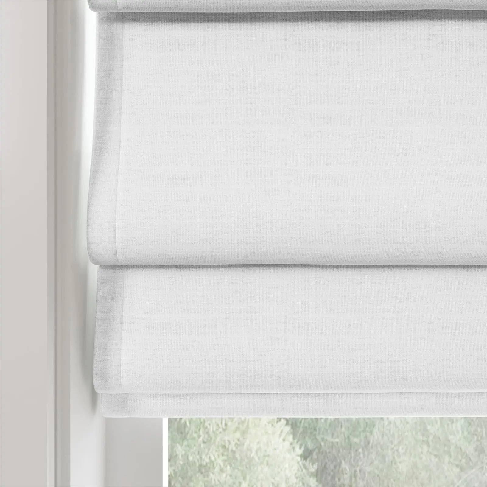 A white linen textured Roman blind with soft stacked folds partially revealing blurred green foliage outside a window