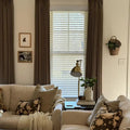 Photograph of a warm and inviting living room interior featuring a beige sectional sofa with patterned floral pillows and a cream knit throw a brass task lamp illuminating books on an end table including 'The Art of Home' and a woven wall basket holding a green plant