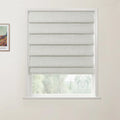 Photograph showing a textured light beige Roman blind with crisp horizontal folds covering a white-framed window, revealing a glimpse of blurred green trees, with a framed Van Gogh landscape print on the adjacent wall