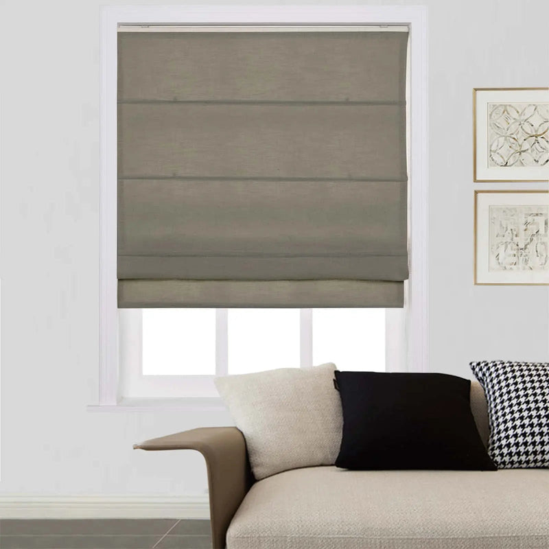 A photograph of a modern living room interior showcasing a window dressed with a medium grey Roman blind, a beige sofa featuring a unique curved armrest, and decorative pillows including a black and white houndstooth pattern, alongside two framed abstract art prints on the wall