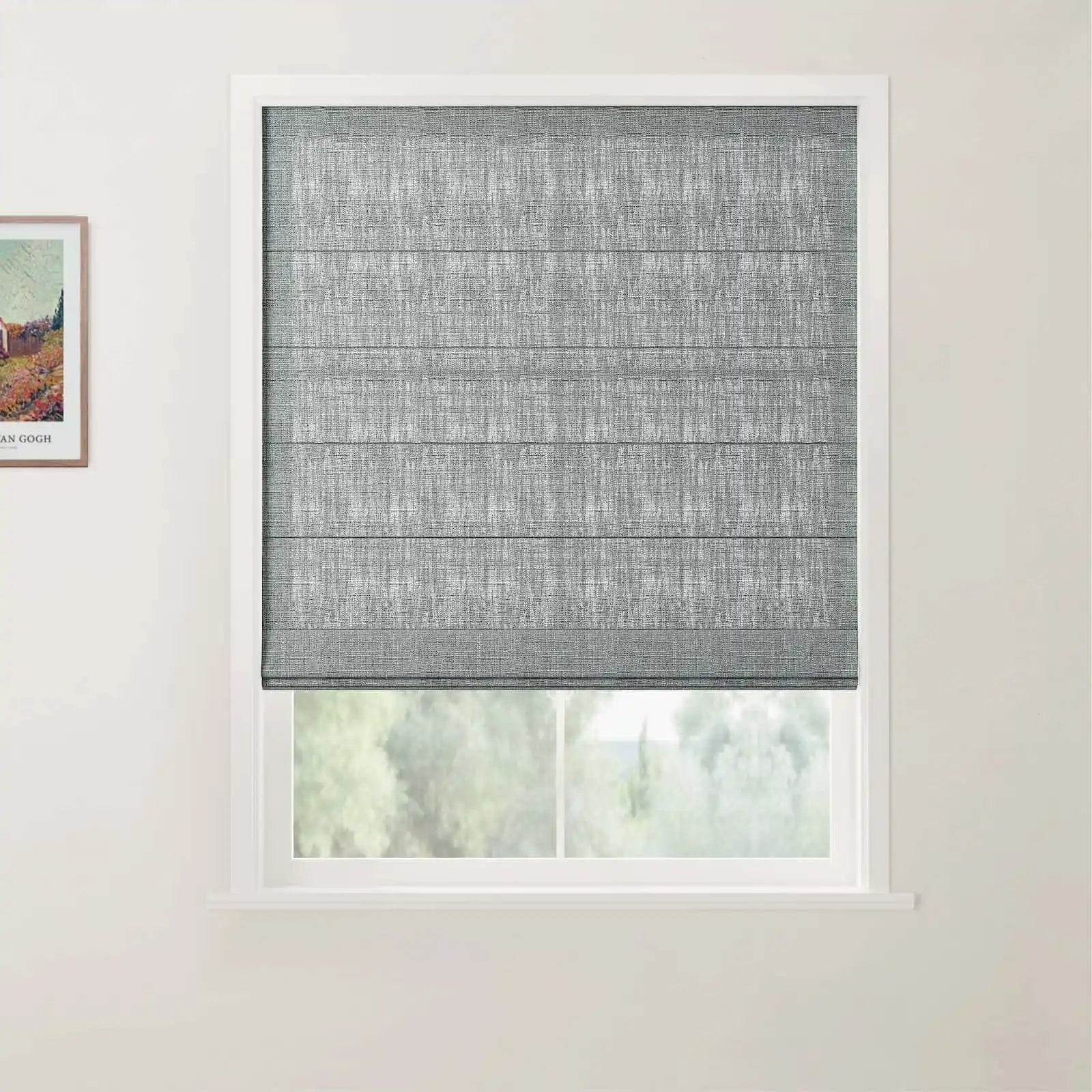 A grey textured Roman blind is partially lowered over a window in a light-colored room revealing a soft green foliage view outside and a framed Van Gogh print on the left wall