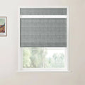 Photograph of a contemporary room featuring a grey linen flat-fold Roman blind covering the top portion of a white-framed window, revealing blurred lush green outdoor foliage below, and a framed Van Gogh landscape print on the adjacent wall