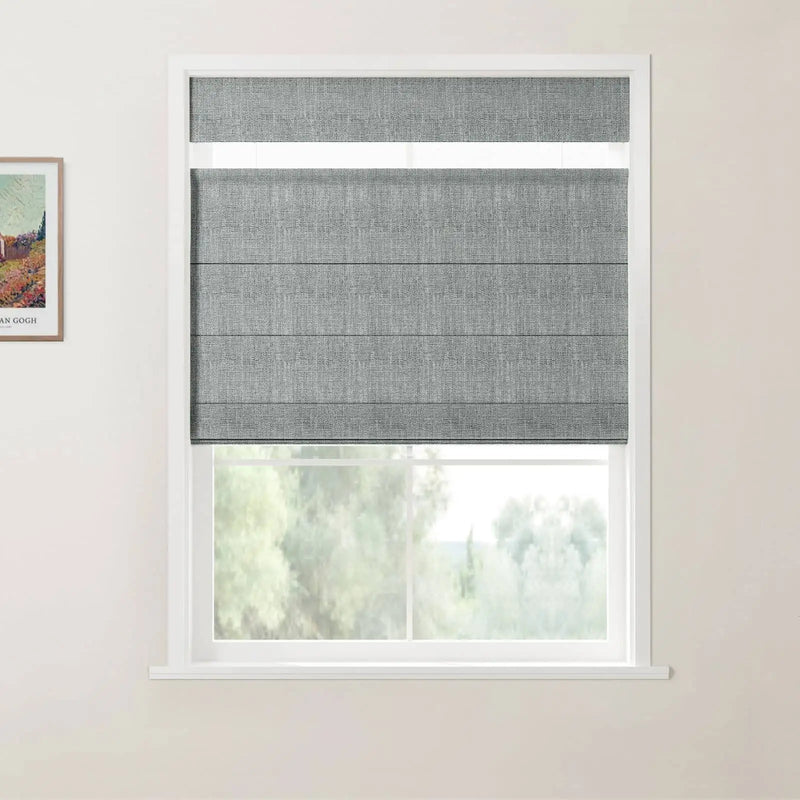 Photograph of a contemporary room featuring a grey linen flat-fold Roman blind covering the top portion of a white-framed window, revealing blurred lush green outdoor foliage below, and a framed Van Gogh landscape print on the adjacent wall