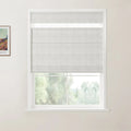 Photograph of a room featuring a window with a light grey textured Roman blind, a white frame, a framed Van Gogh landscape print on the cream wall, and blurred green trees outside
