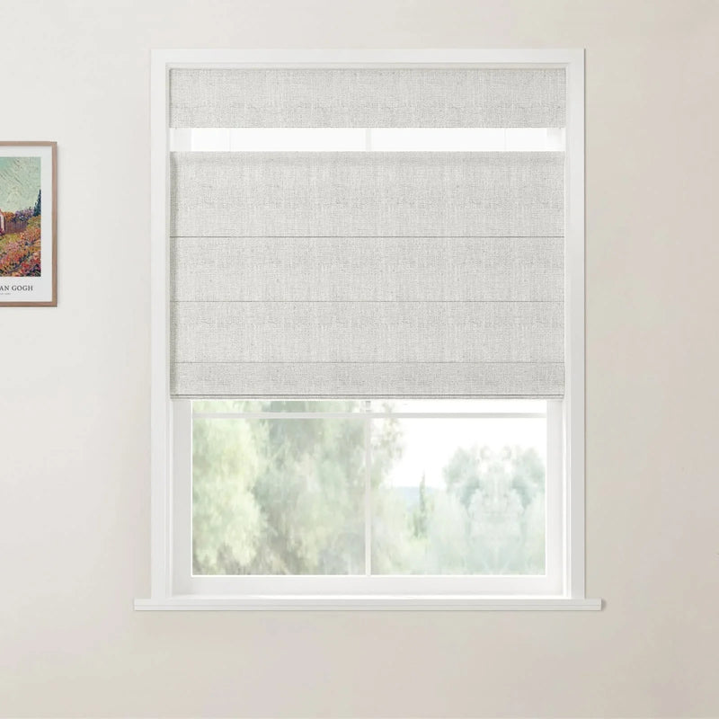 Photograph of a room featuring a window with a light grey textured Roman blind, a white frame, a framed Van Gogh landscape print on the cream wall, and blurred green trees outside