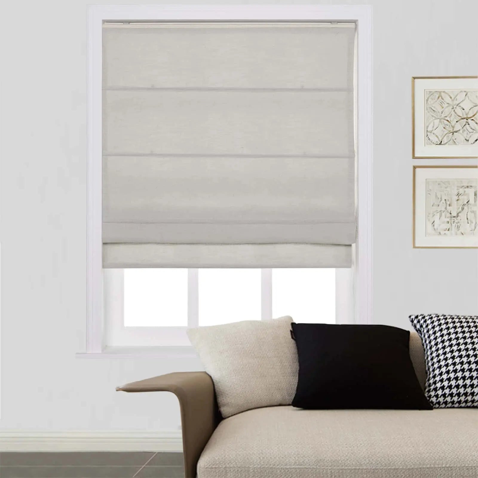 Interior scene showing a window fitted with a light beige Roman blind, an off-white textured sofa with cream, black, and houndstooth pattern cushions, and two gold-framed abstract artworks on the adjacent wall