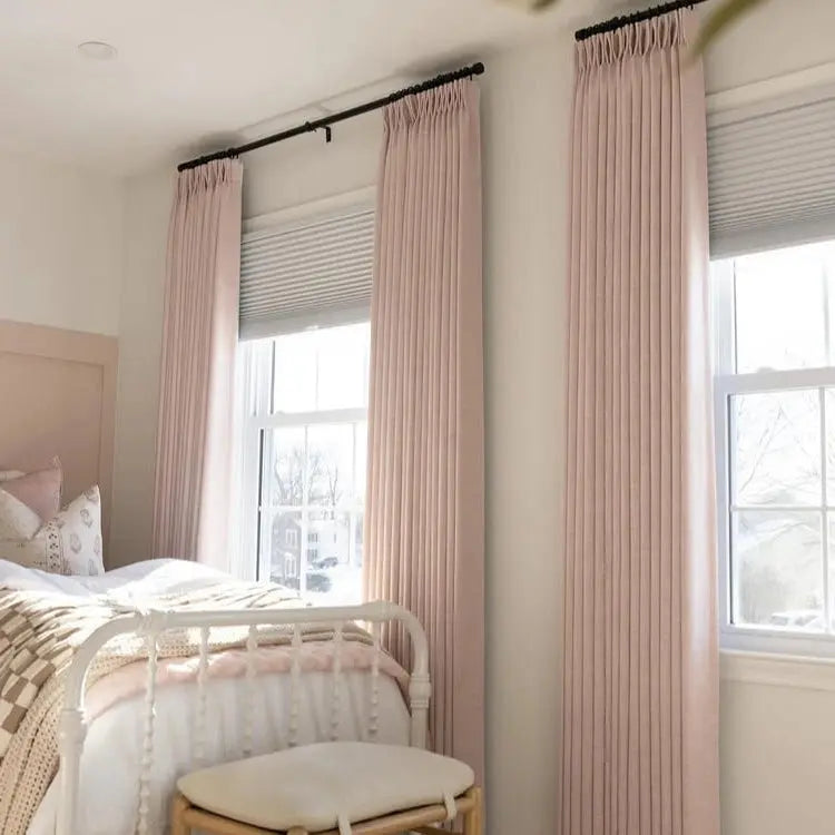 A serene bedroom interior featuring blush pink pleated drapes on black rods, a white metal bed frame, and a woven checkered throw blanket over a light wooden stool