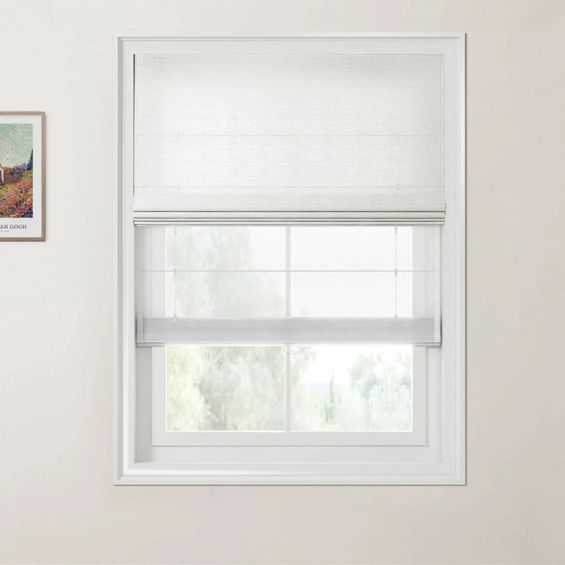 Photograph of a white framed window featuring an opaque white Roman blind above a sheer white roller blind, with green foliage visible outside and a framed Van Gogh print partially on the left wall