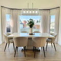 A bright modern dining room interior features a large white rectangular dining table, six beige upholstered chairs, a linear glass chandelier, and a vase of green foliage as a centerpiece, with cream drapes framing bay windows overlooking a suburban backyard and houses