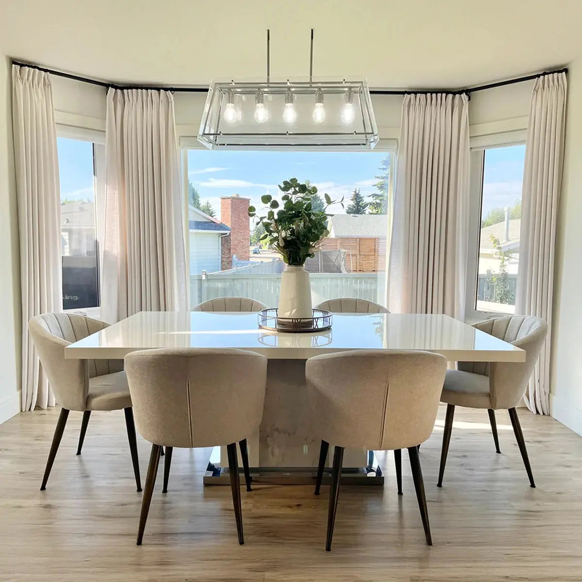 A bright modern dining room interior features a large white rectangular dining table, six beige upholstered chairs, a linear glass chandelier, and a vase of green foliage as a centerpiece, with cream drapes framing bay windows overlooking a suburban backyard and houses