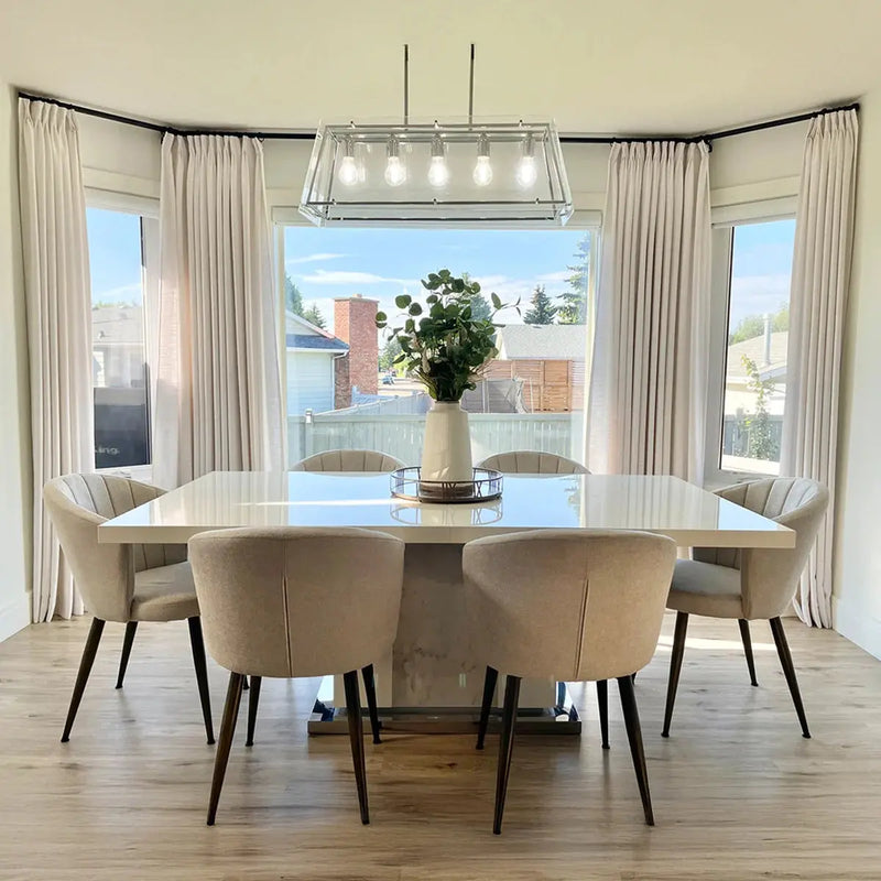 A bright modern dining room interior features a large white rectangular dining table, six beige upholstered chairs, a linear glass chandelier, and a vase of green foliage as a centerpiece, with cream drapes framing bay windows overlooking a suburban backyard and houses