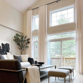 Modern minimalist living room featuring tall floor-to-ceiling windows with white drapes overlooking a lush forest, an abstract black and white painting, and neutral-toned furniture