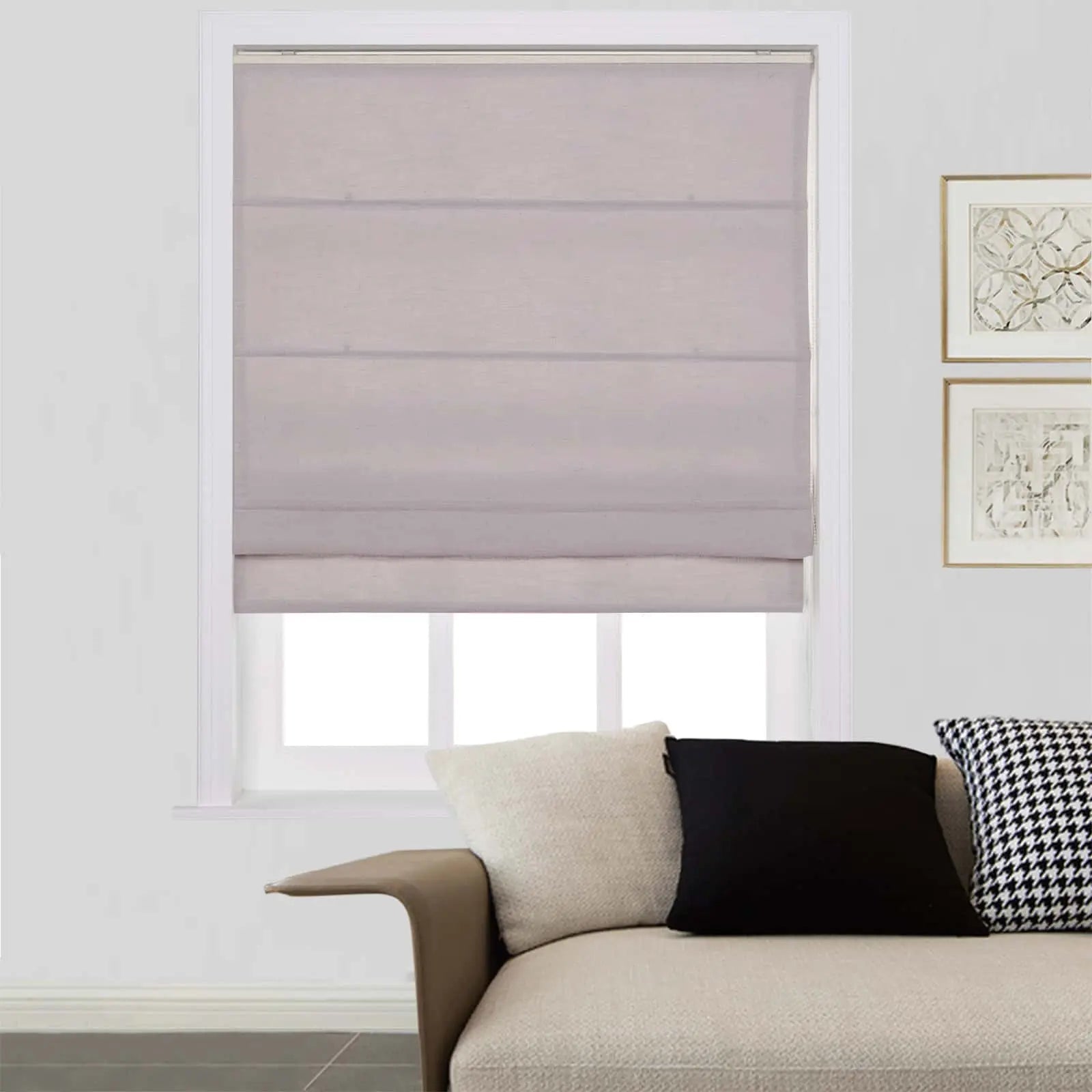 An interior living space featuring a window with a light grey Roman blind, a cream textured sofa adorned with black and houndstooth throw pillows, and two gold-framed abstract artworks