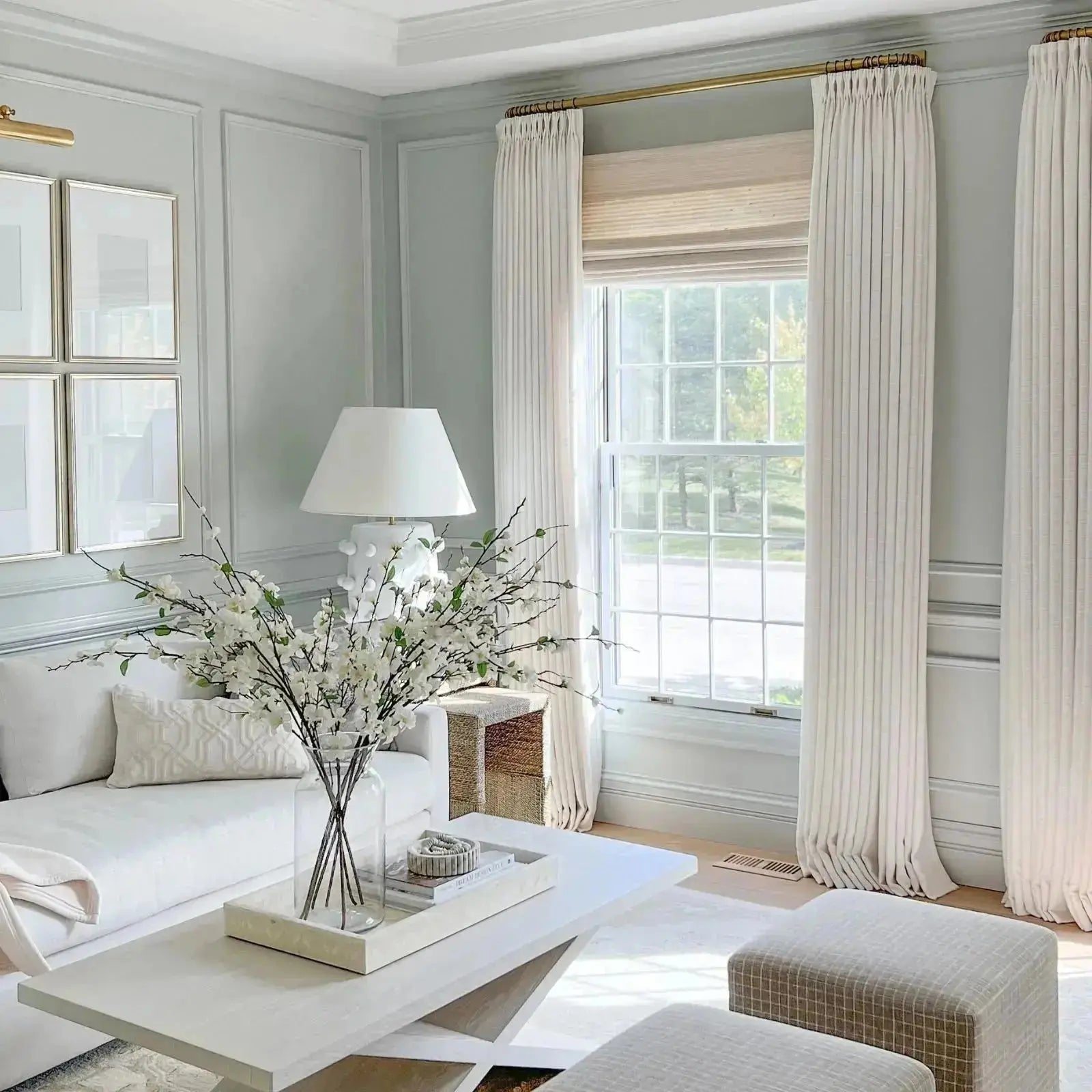 Bright modern living room with a white sofa and coffee table, a vase of white flowering branches, pale gray paneled walls adorned with gold framed art, and a large grid-paned window with elegant ivory drapes on gold rods