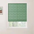 Photograph of a window with a textured sage green Roman blind featuring horizontal folds, set in a white frame against a light beige wall with a framed Van Gogh print to the left