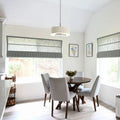 Bright modern kitchen dining nook featuring a round dark wood table with four light gray upholstered chairs, large windows with layered gray roman shades, a white drum pendant light, and two framed blue floral watercolor prints