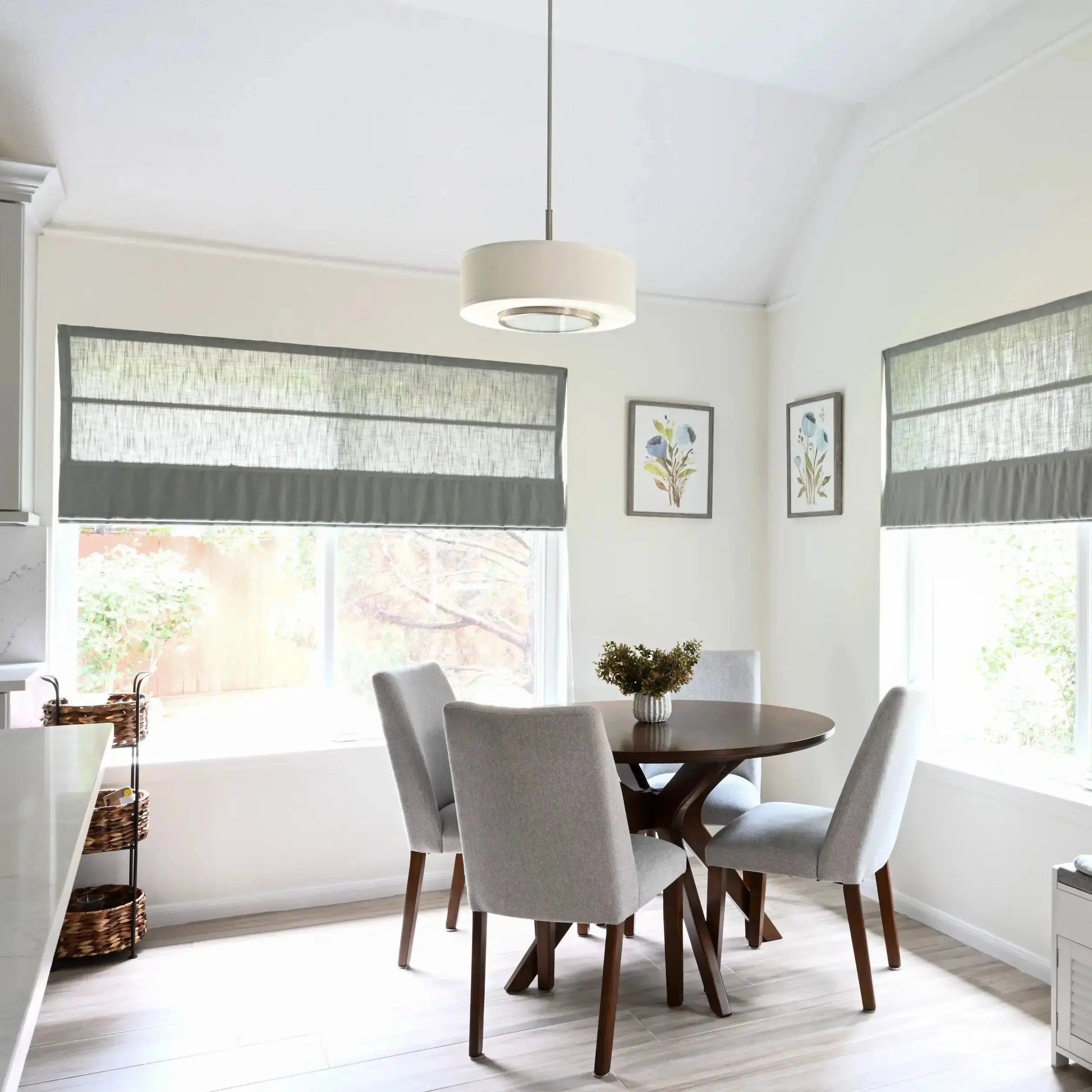 Bright modern kitchen dining nook featuring a round dark wood table with four light gray upholstered chairs, large windows with layered gray roman shades, a white drum pendant light, and two framed blue floral watercolor prints