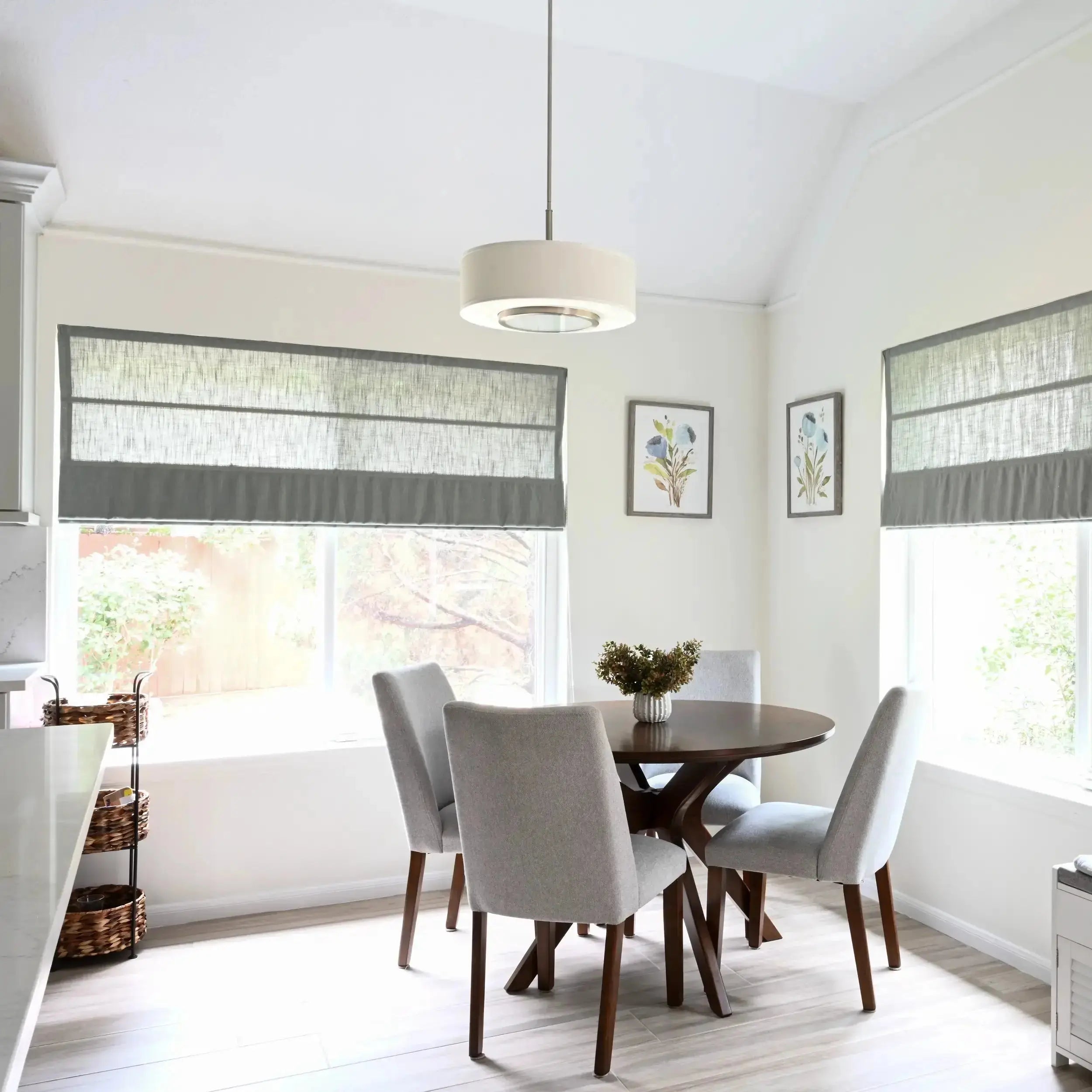 Bright modern kitchen dining nook featuring a round dark wood table with four light gray upholstered chairs, large windows with layered gray roman shades, a white drum pendant light, and two framed blue floral watercolor prints