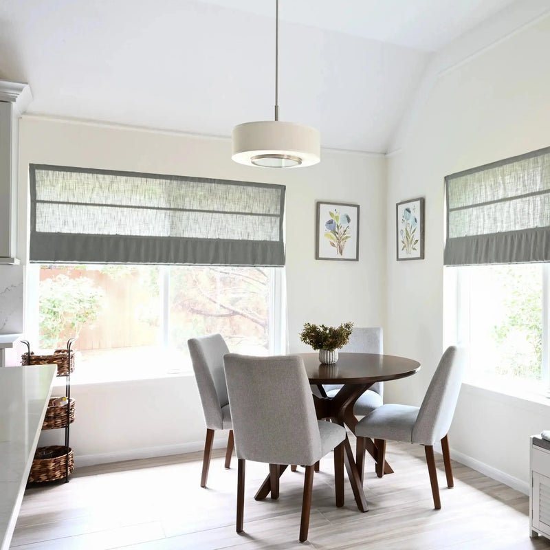 Bright modern kitchen dining nook featuring a round dark wood table with four light gray upholstered chairs, large windows with layered gray roman shades, a white drum pendant light, and two framed blue floral watercolor prints