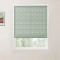 A sage green textured Roman blind with horizontal folds in a white window frame, partially revealing a blurry green landscape outside, with a framed Van Gogh print on the adjacent wall