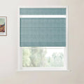Print of Van Gogh's painting on an off-white wall next to a window featuring a partially lowered textured teal roman blind revealing blurry green trees outside