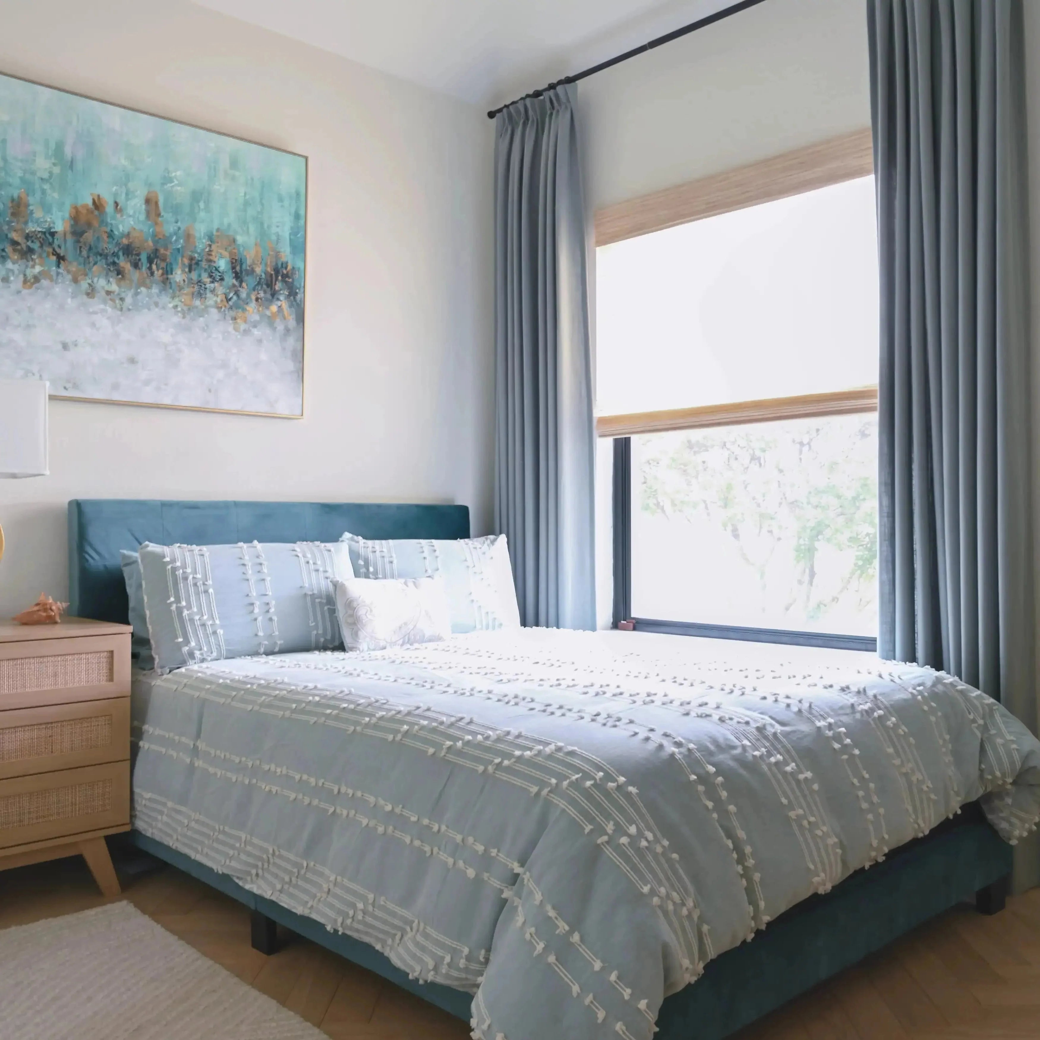 Modern bedroom with a teal velvet bed, light blue textured bedding, large abstract art featuring teal and gold, and a rattan nightstand beside a bright window