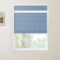 Photograph of a window with a folded blue textured Roman blind covering most of the glass, set in a white frame on a light wall with a framed Van Gogh landscape print visible on the left and blurred green trees beyond the window