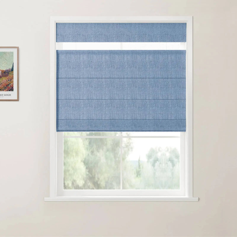 Photograph of a window with a folded blue textured Roman blind covering most of the glass, set in a white frame on a light wall with a framed Van Gogh landscape print visible on the left and blurred green trees beyond the window