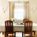 Elegant dining room interior with a white hydrangea centerpiece on a linen tablecloth surrounded by dark wood cane back chairs and brass wall sconces