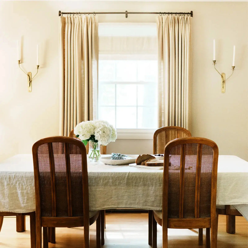 Elegant dining room interior with a white hydrangea centerpiece on a linen tablecloth surrounded by dark wood cane back chairs and brass wall sconces