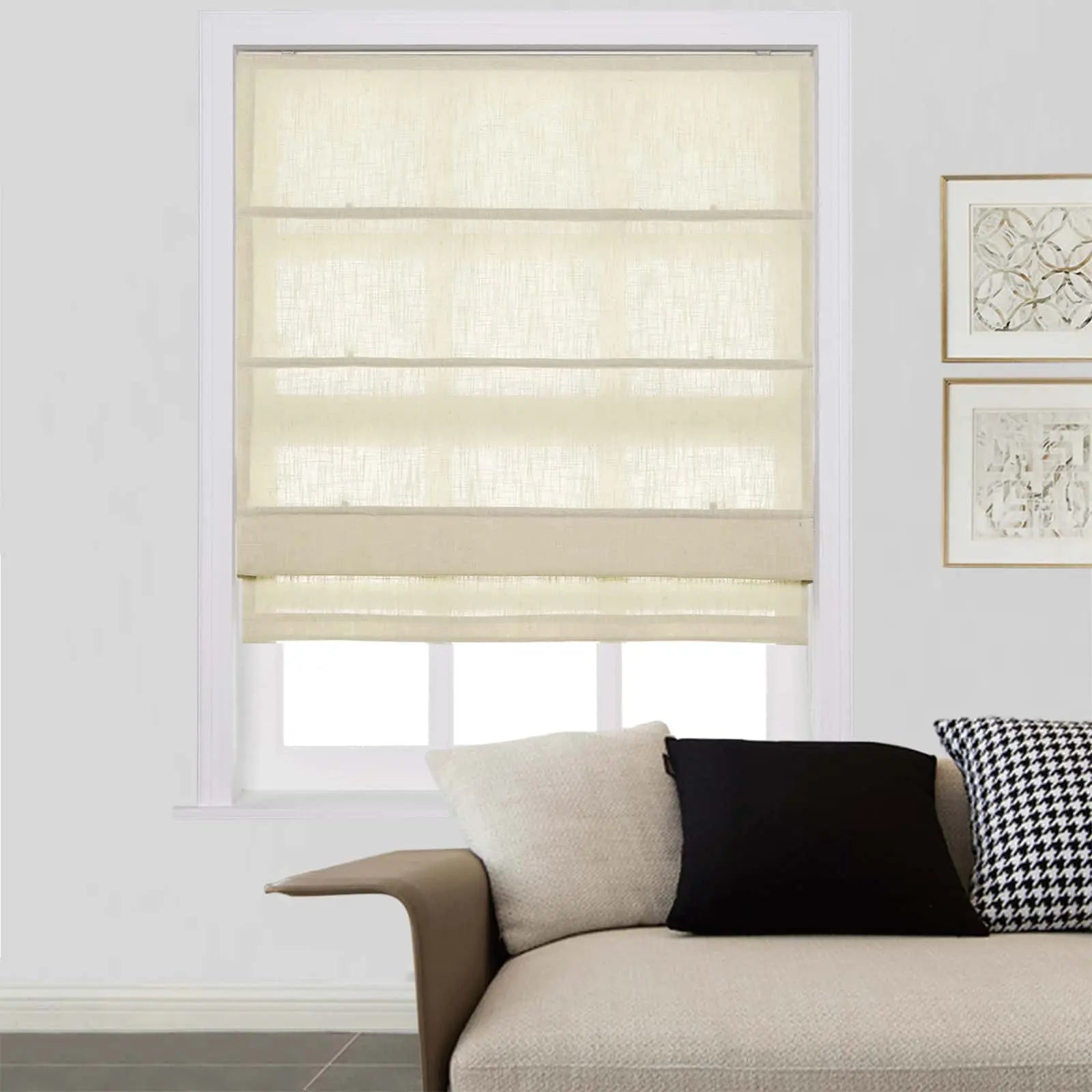 Photograph of a bright window fitted with off-white linen Roman shades, illuminating a modern living room featuring a beige sofa adorned with cream, black, and houndstooth pillows and abstract framed artwork