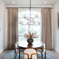 Modern dining room interior with a large arched window framed by beige curtains, a dark oval dining table with white upholstered chairs, a prominent multi-arm black chandelier, and a pampas grass centerpiece