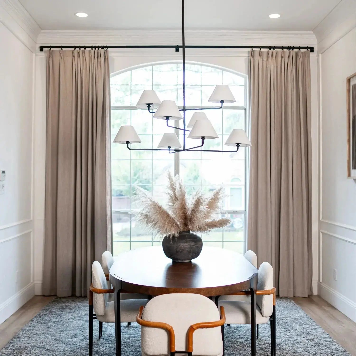 Modern dining room interior with a large arched window framed by beige curtains, a dark oval dining table with white upholstered chairs, a prominent multi-arm black chandelier, and a pampas grass centerpiece