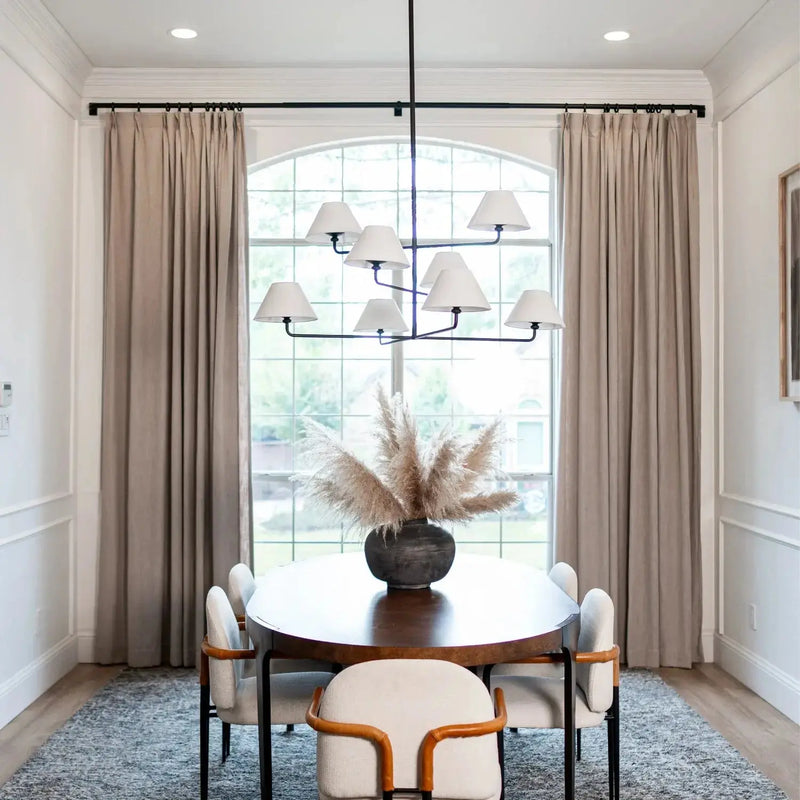 Modern dining room interior with a large arched window framed by beige curtains, a dark oval dining table with white upholstered chairs, a prominent multi-arm black chandelier, and a pampas grass centerpiece