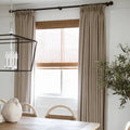 Interior dining room featuring a window with layered beige linen curtains and woven bamboo blinds, a black geometric chandelier, and a light wooden dining table with woven-back chairs and olive branches