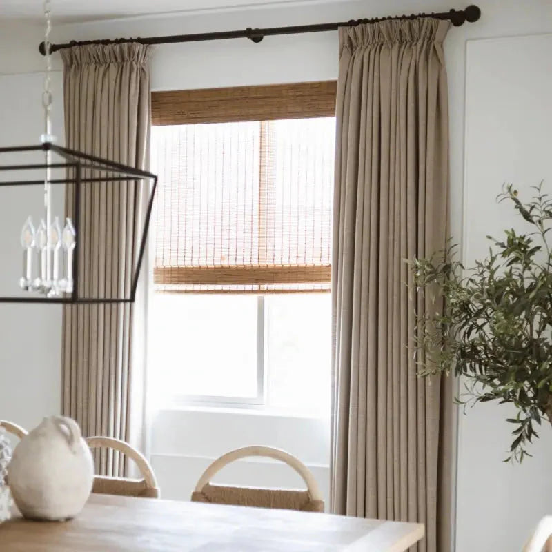 Interior dining room featuring a window with layered beige linen curtains and woven bamboo blinds, a black geometric chandelier, and a light wooden dining table with woven-back chairs and olive branches