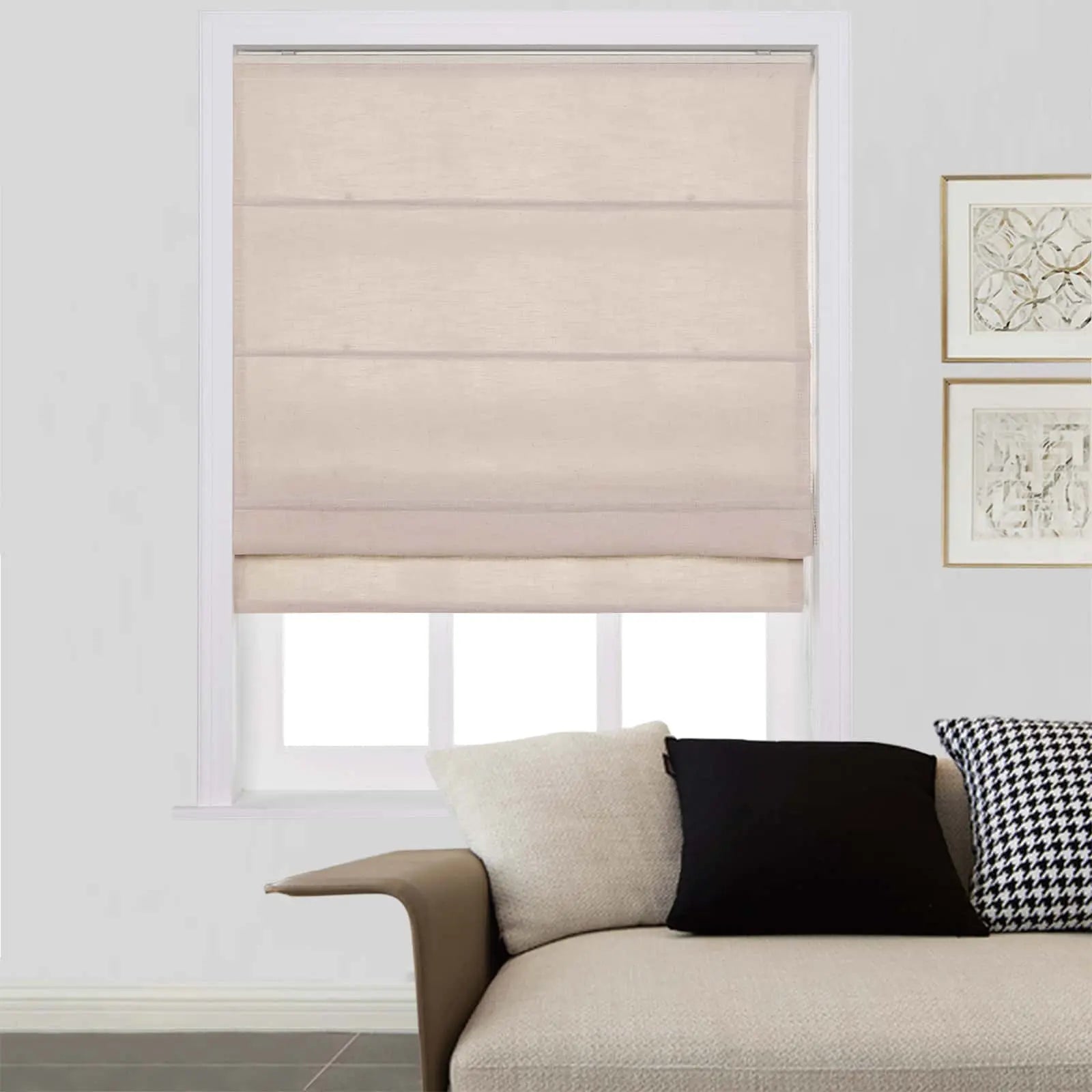 A modern living room interior showcasing a window with a light beige Roman blind, a textured cream sofa with a black and houndstooth pillow, and two framed abstract artworks on the wall