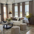 A warm and inviting modern living room featuring a plush white sectional sofa, floor-to-ceiling taupe drapes, a dark wood coffee table with fresh greenery, and a distinctive black sputnik chandelier