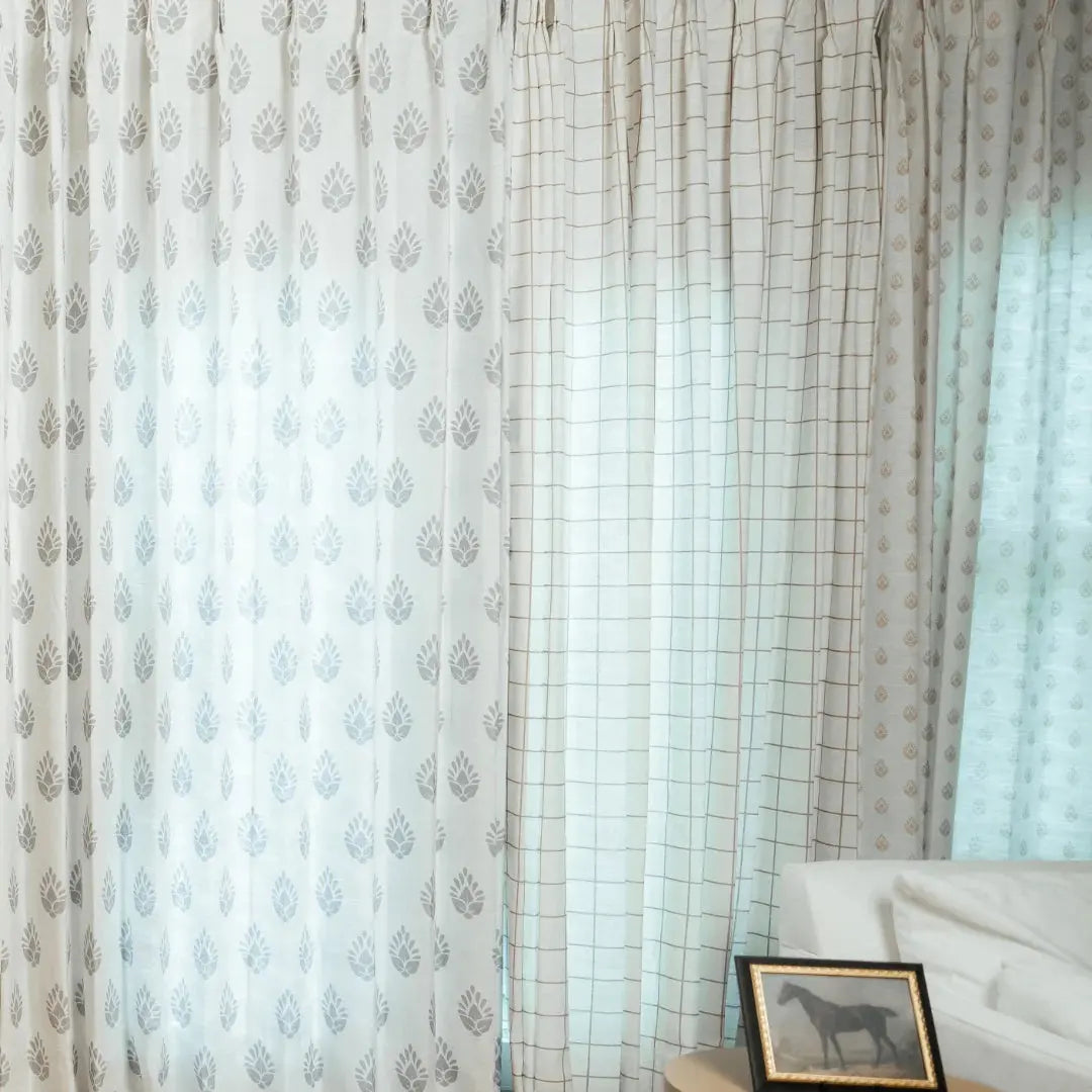 Photograph of an interior window with white curtains showcasing grey leaf patterns, a sheer beige plaid curtain, and a gold-framed horse print on a side table