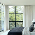 A modern bedroom interior with large black-framed windows showcasing a lush green forest view, elegant white drapes, and a bed adorned with dark gray and white textured quilted comforters