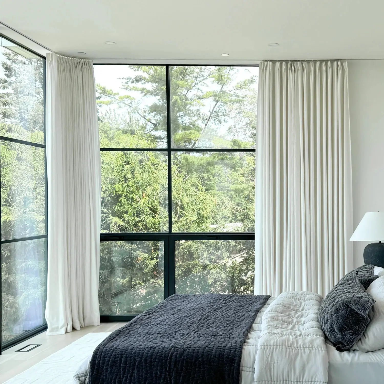 A modern bedroom interior with large black-framed windows showcasing a lush green forest view, elegant white drapes, and a bed adorned with dark gray and white textured quilted comforters