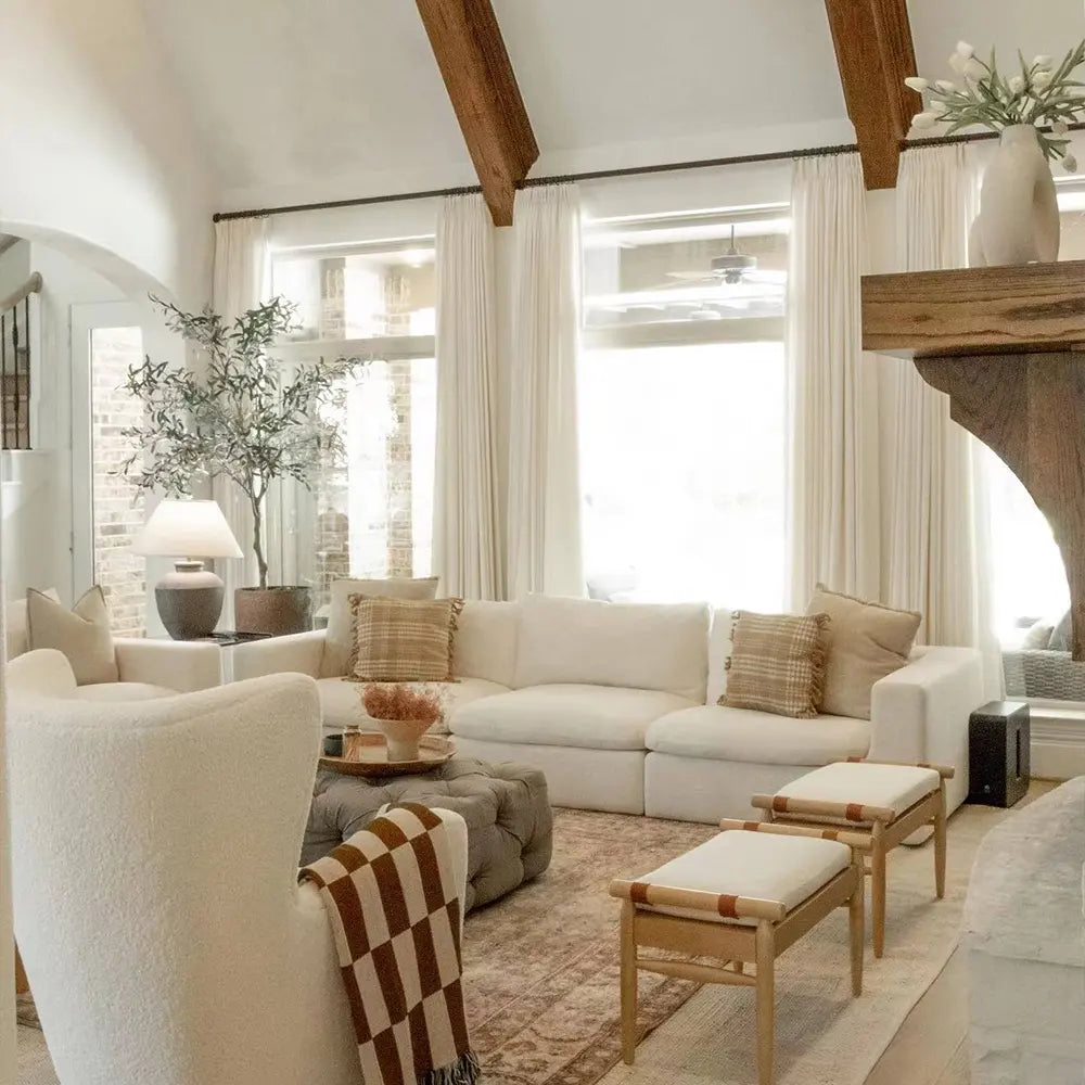 A bright modern farmhouse living room featuring a white modular sofa, exposed dark wood ceiling beams, a large potted olive tree, a tufted grey ottoman, and a brown and white checkered throw blanket draped over an armchair