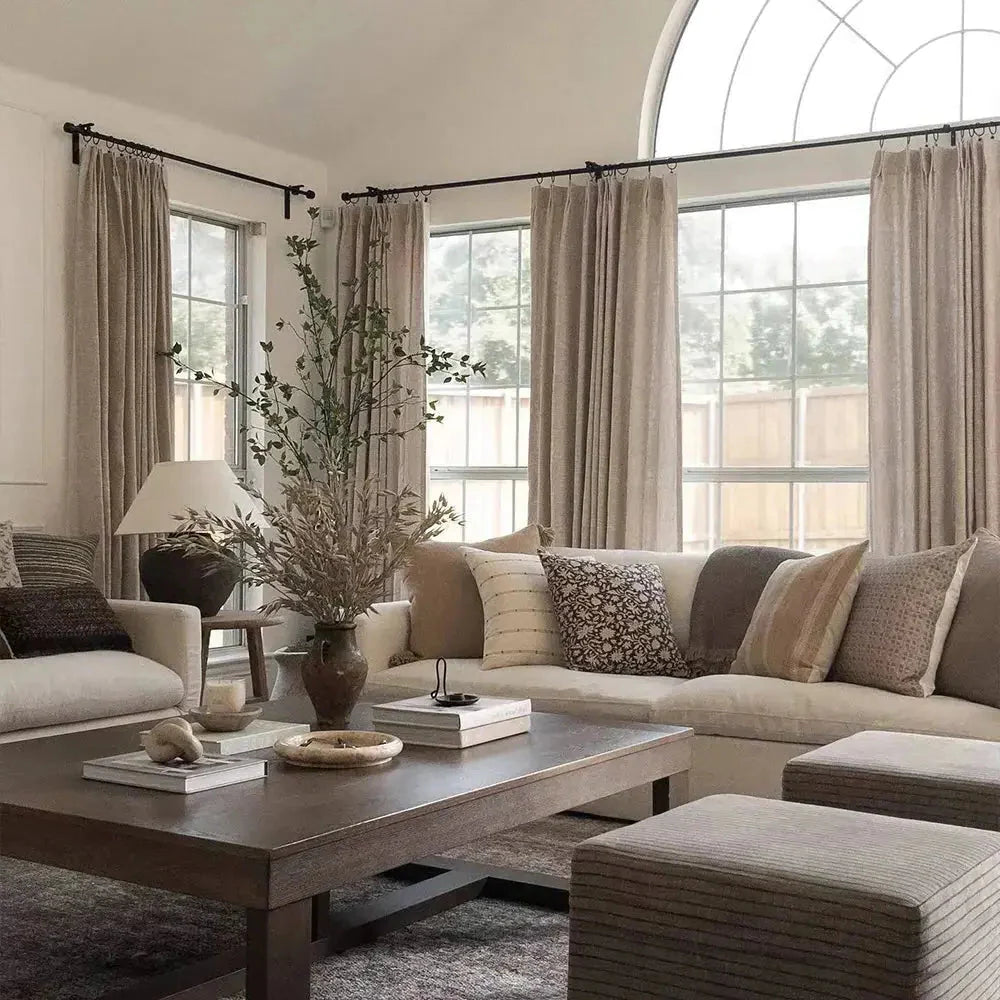 A warm neutral living room interior features a prominent arched window, a cream sofa adorned with various textured and patterned pillows, and a dark wood coffee table displaying a vase of dried and green branches