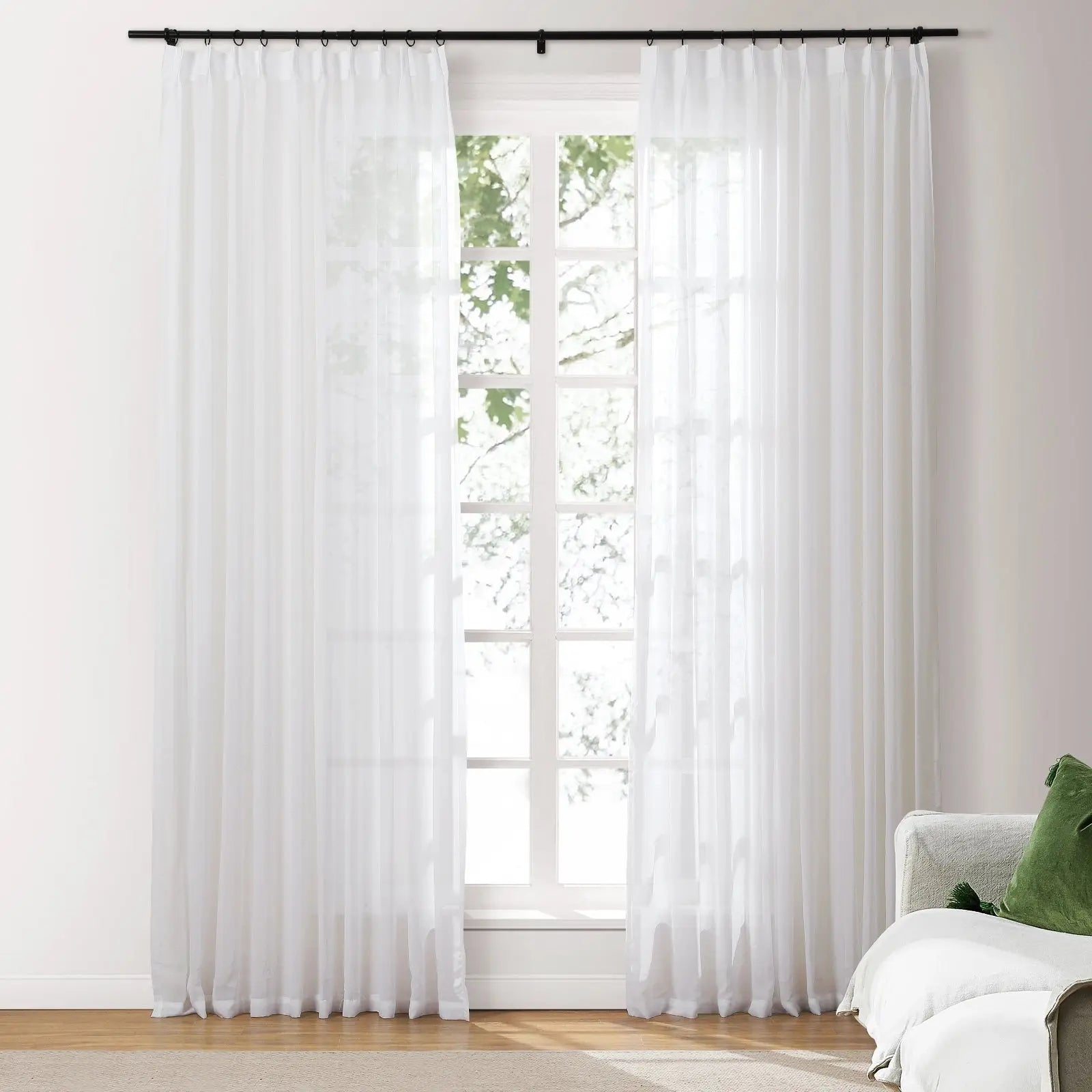 White sheer curtains on a black rod flank a multi-pane window revealing green foliage outside, with a beige sofa and green decorative pillow to the right