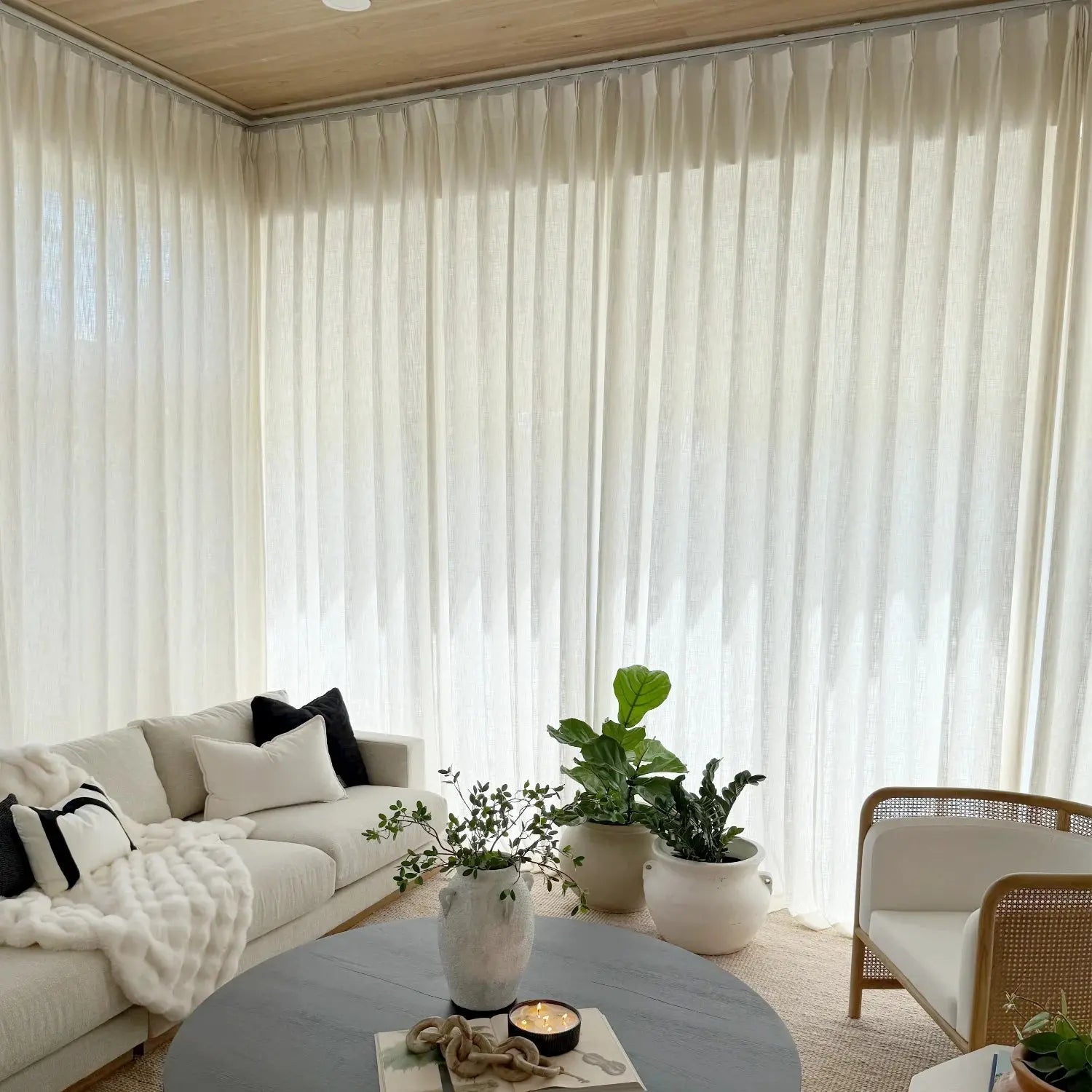A bright modern living room interior with floor-to-ceiling sheer white curtains a white sectional sofa featuring black and white pillows and a round grey coffee table adorned with potted plants and a glowing candle